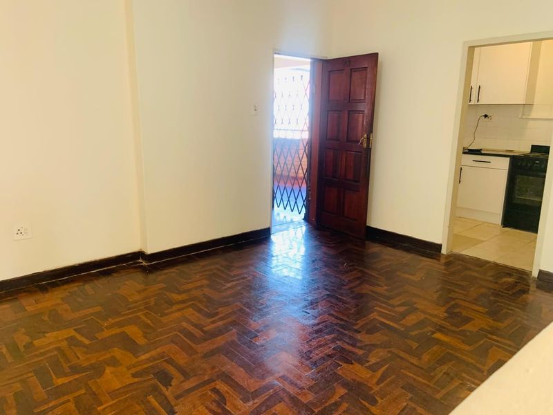 An empty living area featuring classic herringbone parquet flooring, leading to an external security gate and an adjacent kitchen space.