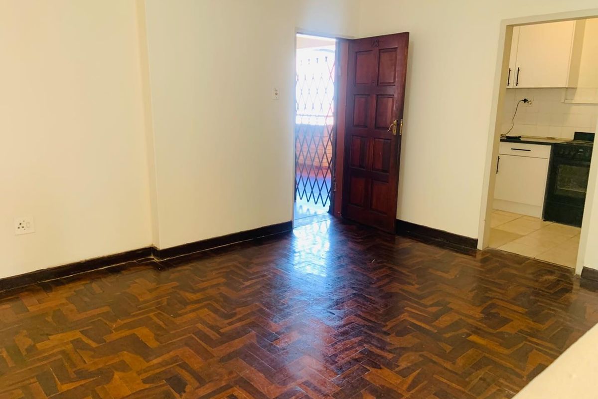 An empty living area featuring classic herringbone parquet flooring, leading to an external security gate and an adjacent kitchen space.