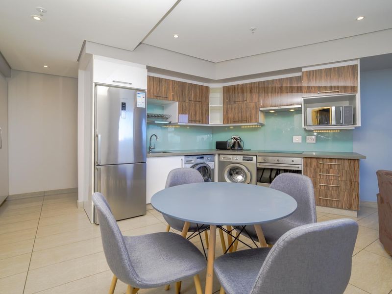 Modern open-plan kitchen featuring wood-grain cabinetry, glass splashback, integrated appliances, and tiled flooring, seamlessly flowing into the living area.