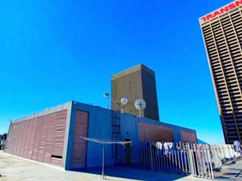 A rooftop terrace area featuring concrete flooring, metal security fencing, and utility structures, offering an open-air space with views of the surrounding city skyline.