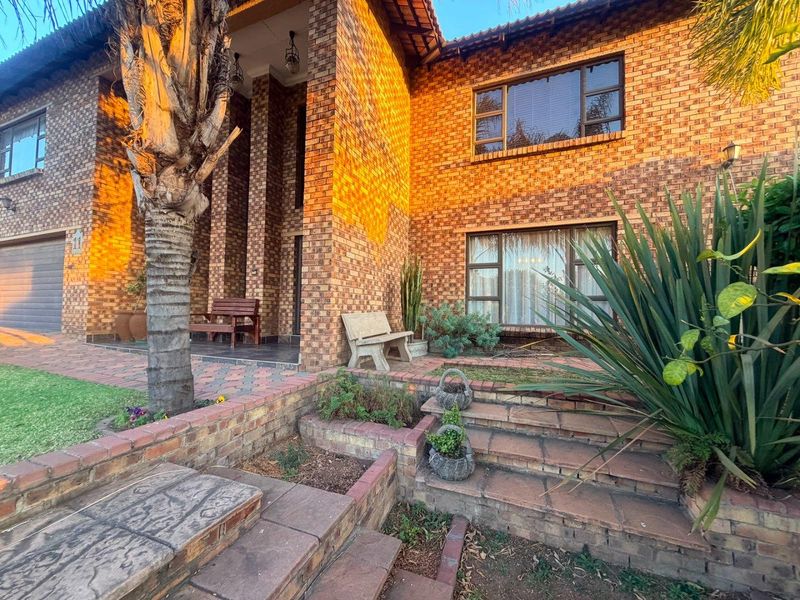 A well-maintained double-storey face-brick home featuring a paved front entrance, established garden landscaping, and secure garage access.