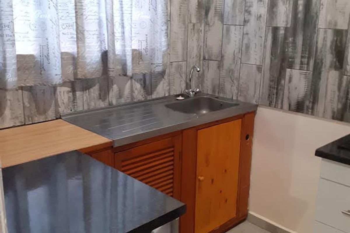 A compact kitchenette area featuring a stainless steel sink, wooden cabinetry, and a tiled backsplash with a modern grey finish.