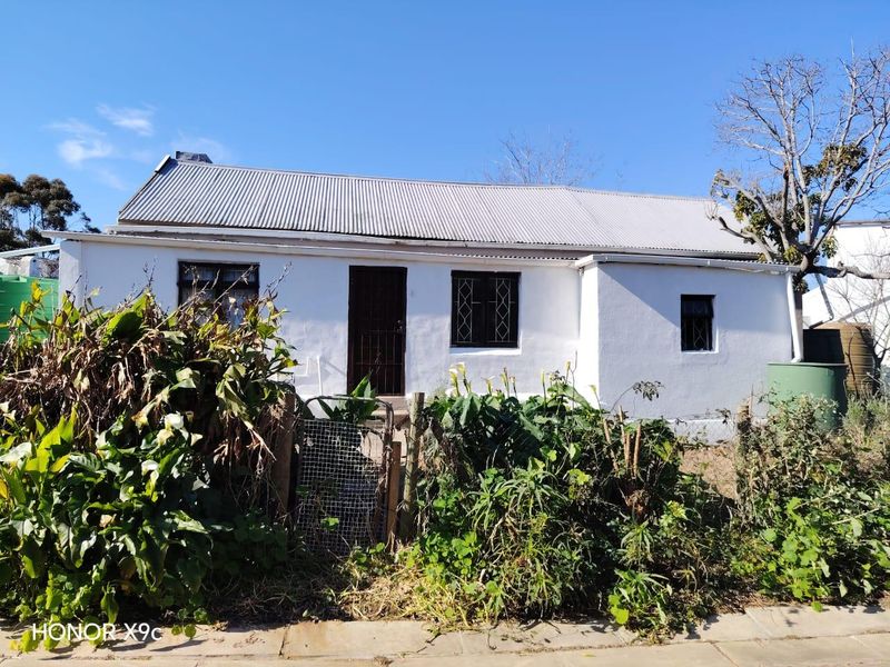 Front elevation of a single-story cottage featuring a corrugated metal roof, security gates, and window bars, set behind overgrown garden foliage.