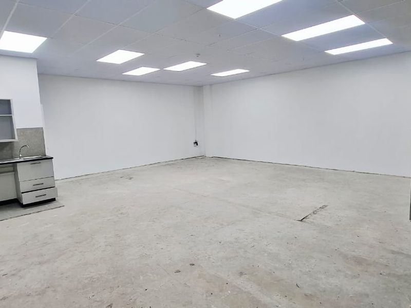 An empty, versatile utility space featuring a suspended ceiling, recessed lighting, a small kitchenette with a sink, and a glass entrance door.