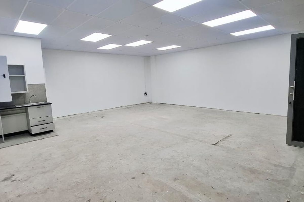 An empty, versatile utility space featuring a suspended ceiling, recessed lighting, a small kitchenette with a sink, and a glass entrance door.
