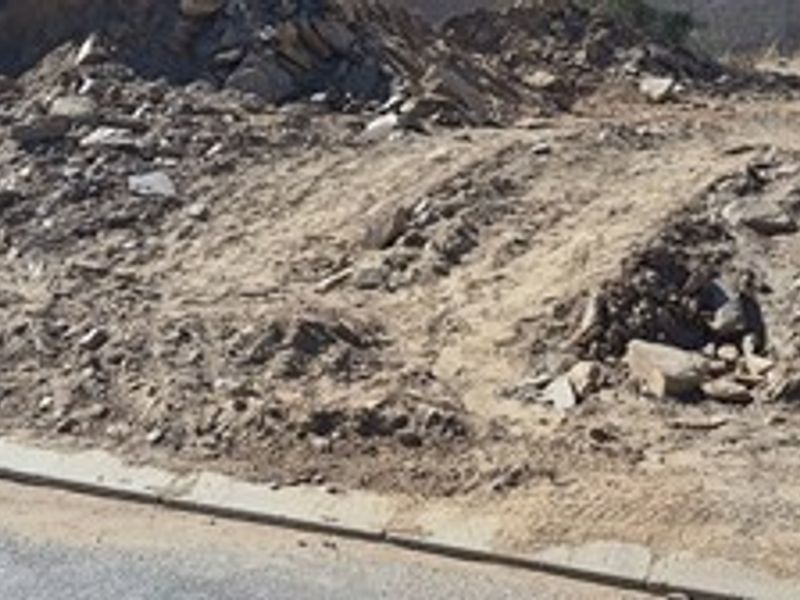 A view of a vacant residential plot showing uneven ground, rocky terrain, and a concrete curb along the street edge.