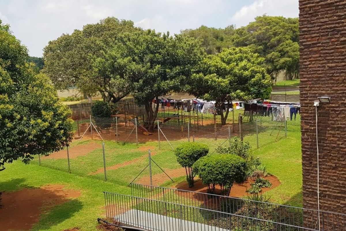 View of the communal garden area featuring established trees, lawn, and security fencing, adjacent to the brick apartment building exterior.
