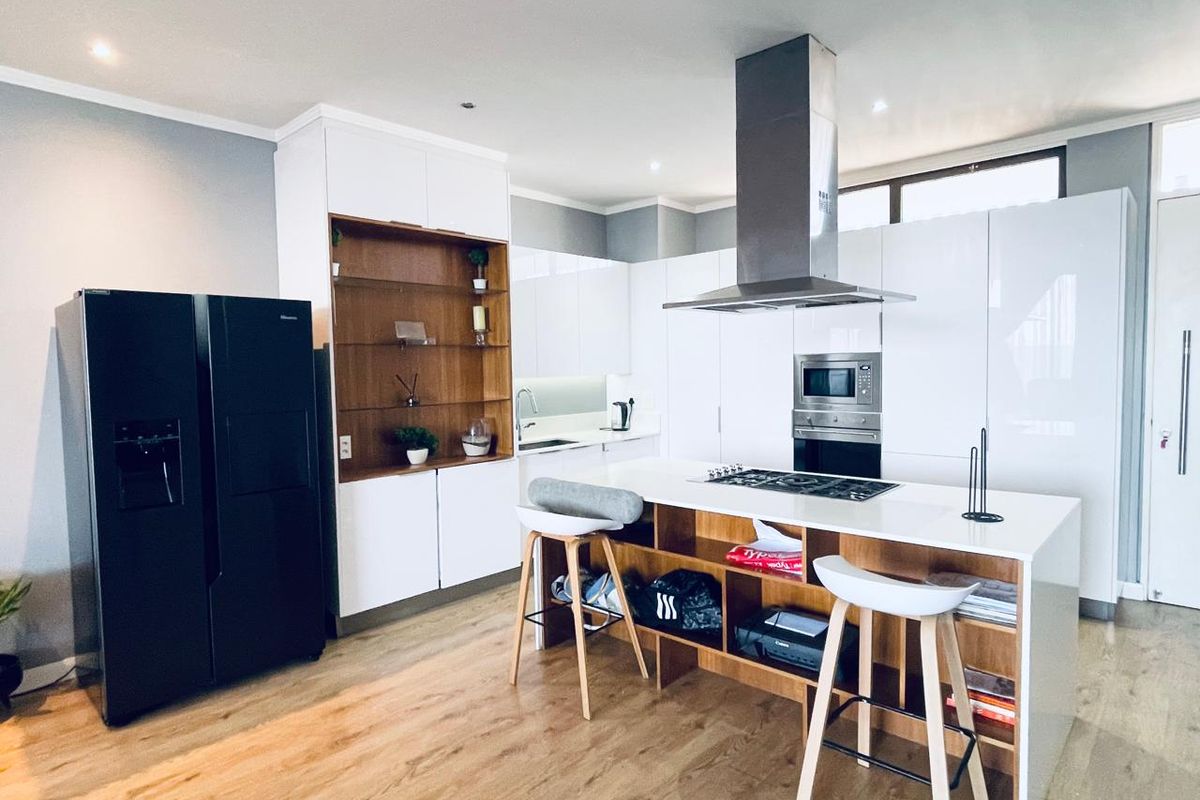 Modern kitchen featuring high-gloss white cabinetry, a central island with a gas hob, integrated appliances, and warm wood-toned shelving and flooring.