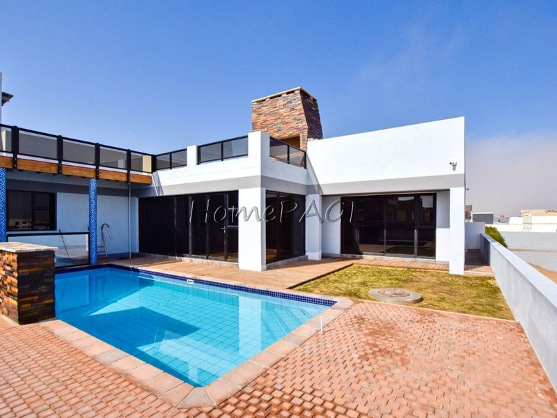 Modern outdoor entertainment area featuring a swimming pool, brick-paved patio, and a distinctive stone-clad chimney, complemented by a balcony with glass balustrades.