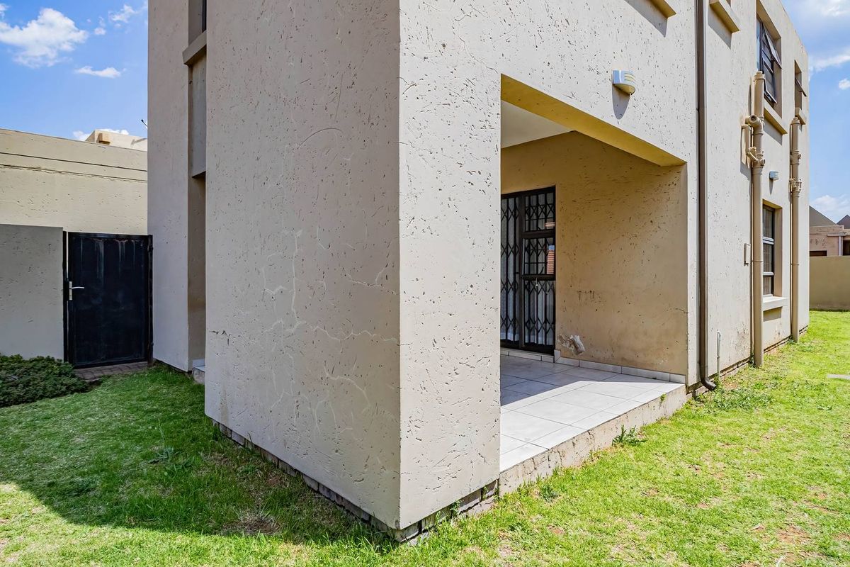 Rear view of the cluster property featuring a small tiled patio, security gate, and surrounding lawn area with boundary walls.