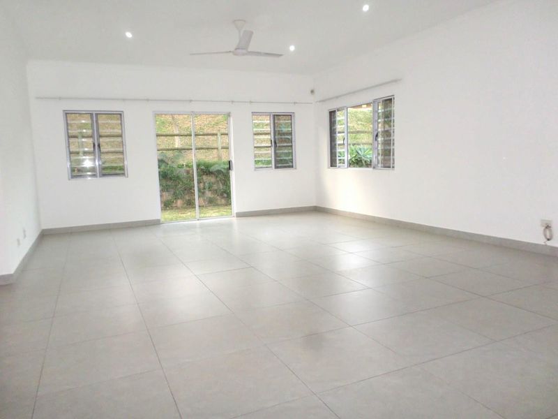 A spacious, bright, and empty living area featuring neutral tiled flooring, recessed lighting, a ceiling fan, and large aluminium windows and sliding doors.