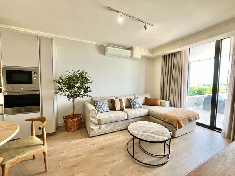 Modern open-plan living area featuring light wood-look flooring, integrated kitchen appliances, split-system air conditioning, and large sliding doors leading to a private balcony.