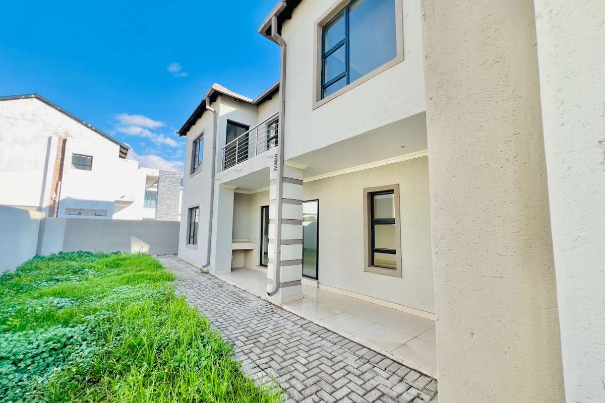 Rear view of a modern two-story house featuring a paved patio area, built-in braai, balcony, and a spacious garden.