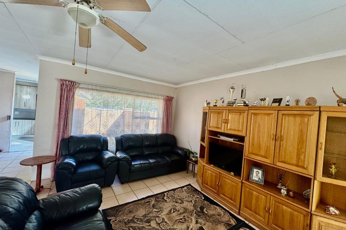 A living room featuring tiled flooring, a ceiling fan, and large windows allowing natural light, with an adjacent view into the kitchen area.