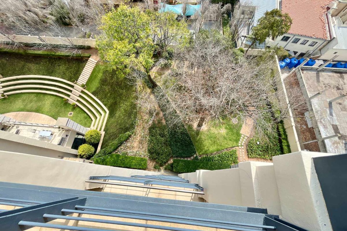 High-angle aerial view overlooking the apartment complex garden, featuring landscaped lawns, terraced retaining walls, and mature trees.