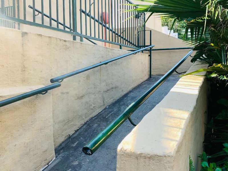 An exterior concrete access ramp featuring metal handrails and security fencing, showing signs of wear and weathering on the surfaces.