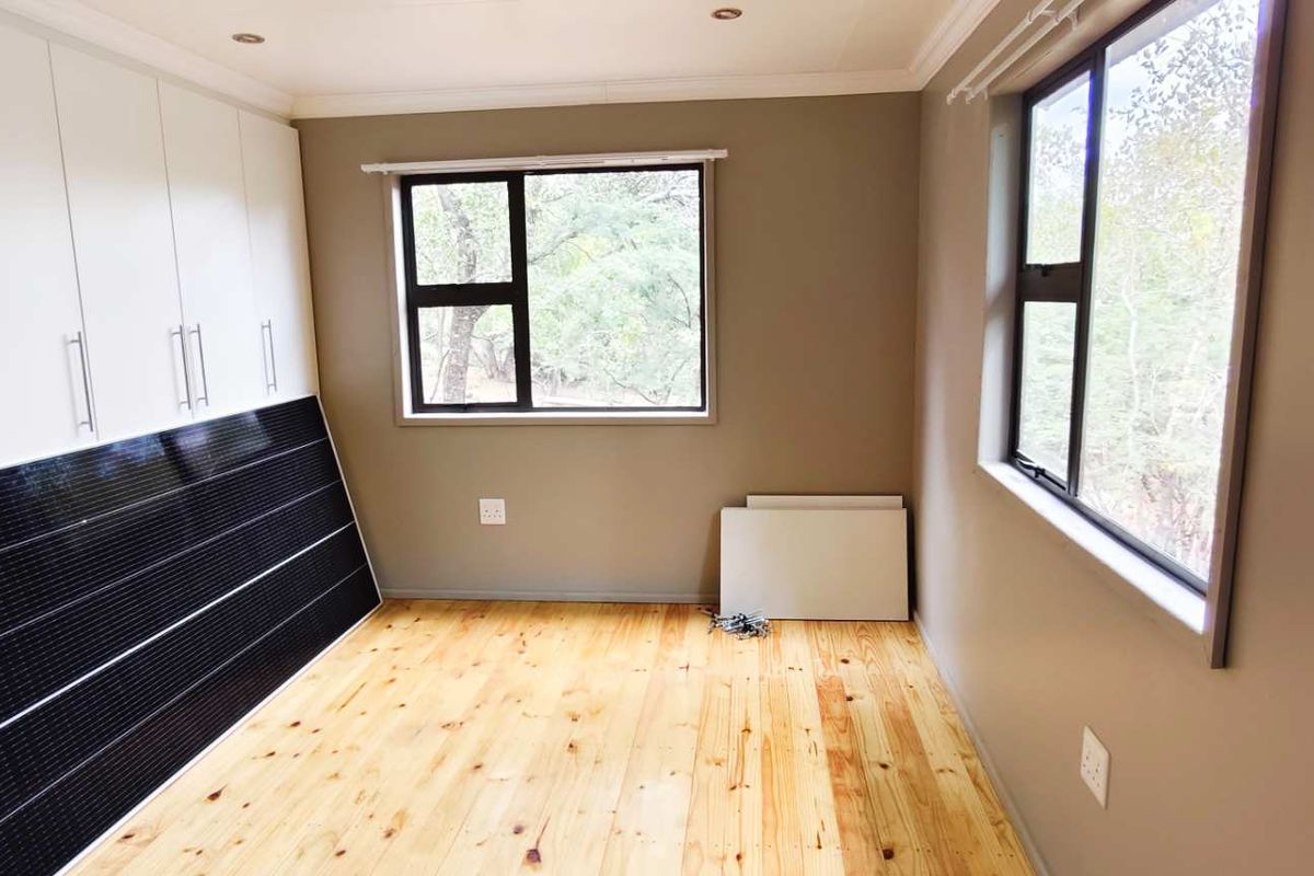 A bright bedroom featuring polished timber flooring, built-in cabinetry, and large windows providing natural light and garden views.