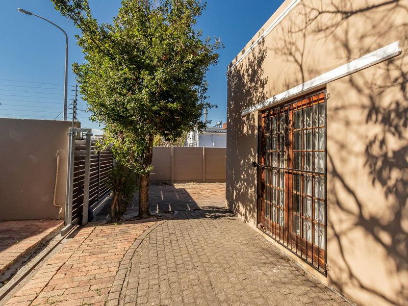 A paved rear exterior area featuring a perimeter wall, mature tree, and wooden french doors secured with a metal gate.
