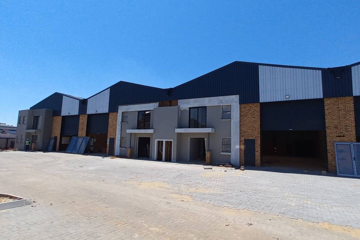 A modern industrial warehouse complex featuring multiple roller shutter doors, metal cladding, and brick facade elements with an expansive paved yard area.