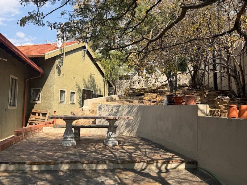 An outdoor patio area featuring stone paving and a retaining wall, shaded by mature trees, suitable for an office break or outdoor workspace.