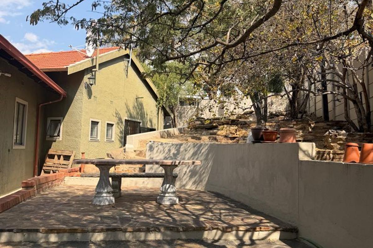 An outdoor patio area featuring stone paving and a retaining wall, shaded by mature trees, suitable for an office break or outdoor workspace.