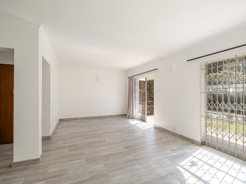 A bright, empty living room featuring modern grey tiled flooring, clean white walls, and a sliding glass door with a security gate leading outside.