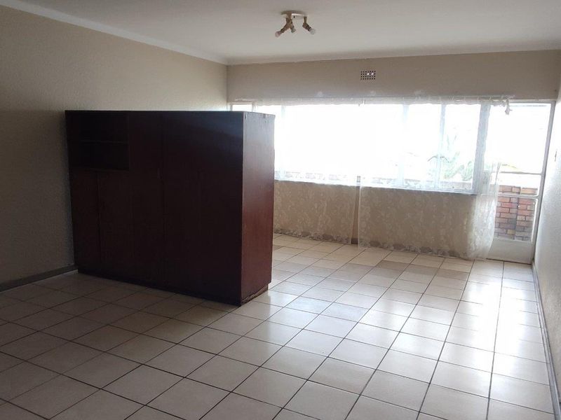 A spacious, empty living area featuring neutral tiled flooring, large windows providing natural light, and a simple ceiling light fixture.