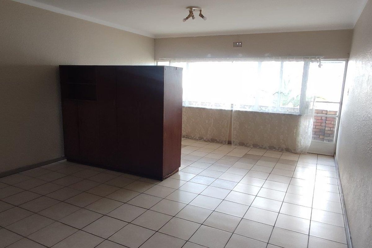 A spacious, empty living area featuring neutral tiled flooring, large windows providing natural light, and a simple ceiling light fixture.