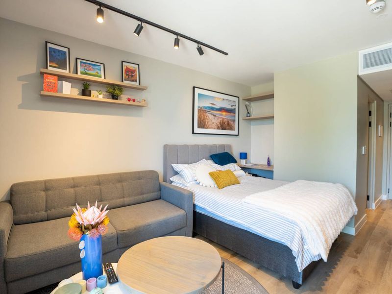 A modern open-plan studio apartment featuring timber flooring, track lighting, built-in shelving, and integrated ducted air conditioning.