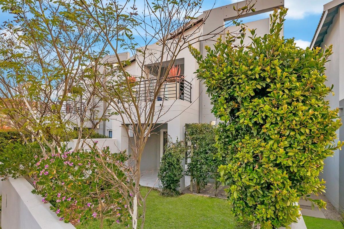 Rear view of the apartment building featuring a private balcony, paved patio area, and a well-maintained landscaped garden.
