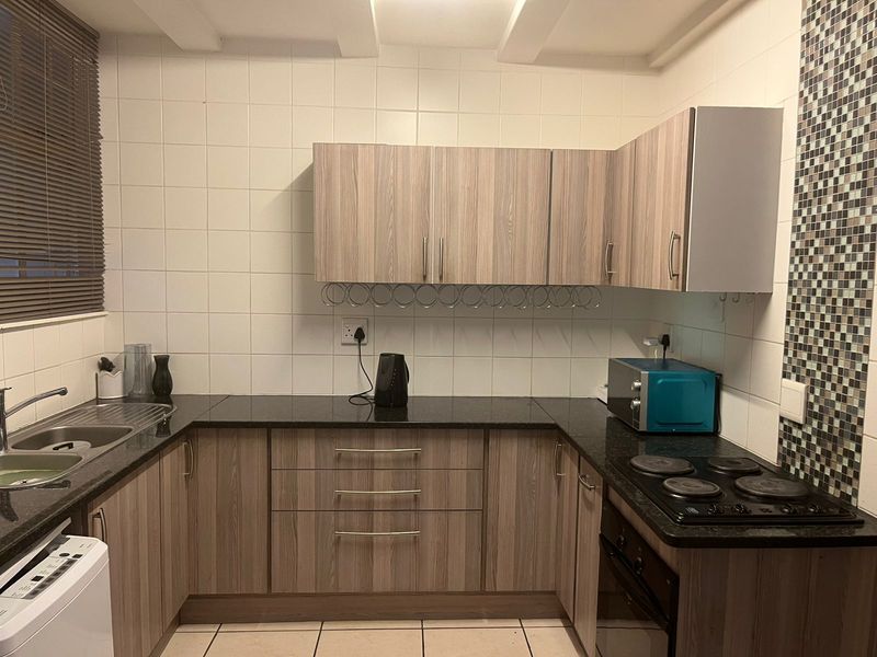 A functional kitchen featuring wood-look laminate cabinetry, granite countertops, a built-in electric hob and oven, and a decorative mosaic tile feature wall.