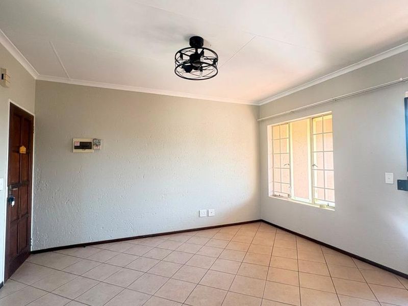 A bright, empty living room featuring neutral tiled flooring, a modern ceiling fan, and a window with security bars.