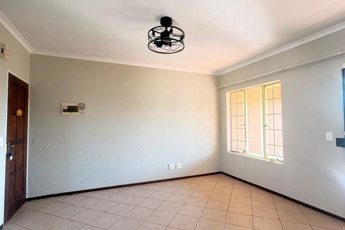 A bright, empty living room featuring neutral tiled flooring, a modern ceiling fan, and a window with security bars.