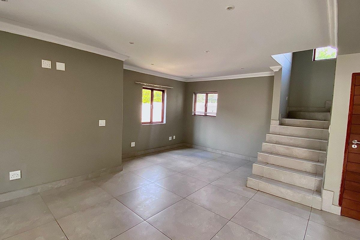 An open-plan living area featuring neutral wall tones, large floor tiles, recessed lighting, and a staircase leading to the upper level.