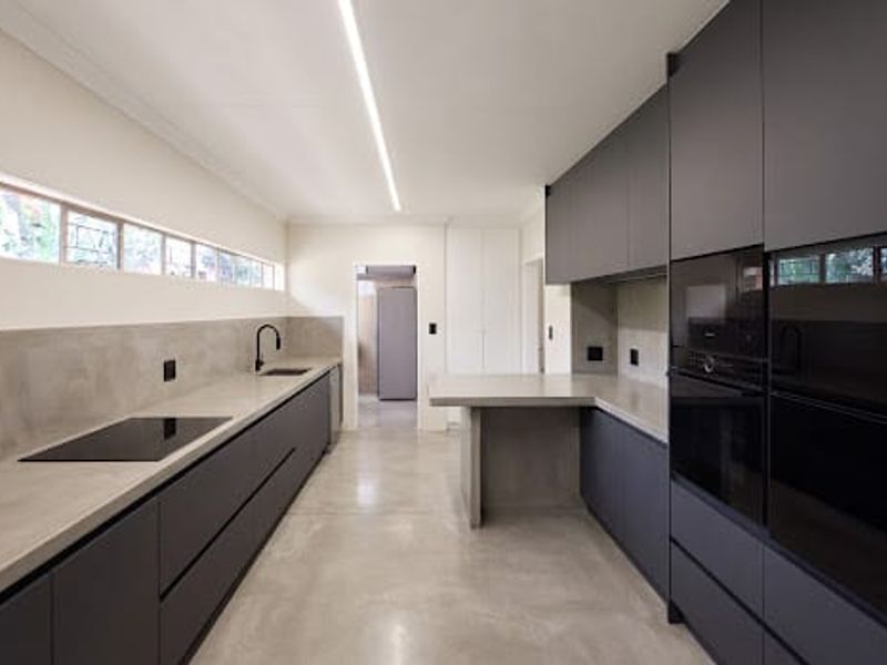 Modern minimalist kitchen featuring matte dark cabinetry, concrete countertops, integrated appliances, and polished concrete floors with sleek linear lighting.