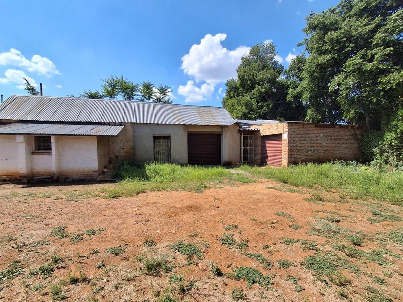 Rear view of a utilitarian building featuring corrugated metal roofing, roller shutter doors, and security gates, set on an unpaved lot with overgrown vegetation.
