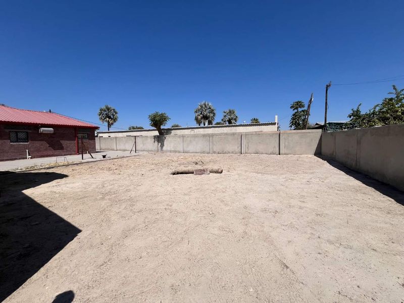 A spacious, undeveloped rear yard featuring a precast concrete boundary wall and views of the adjacent brick building with a red metal roof.