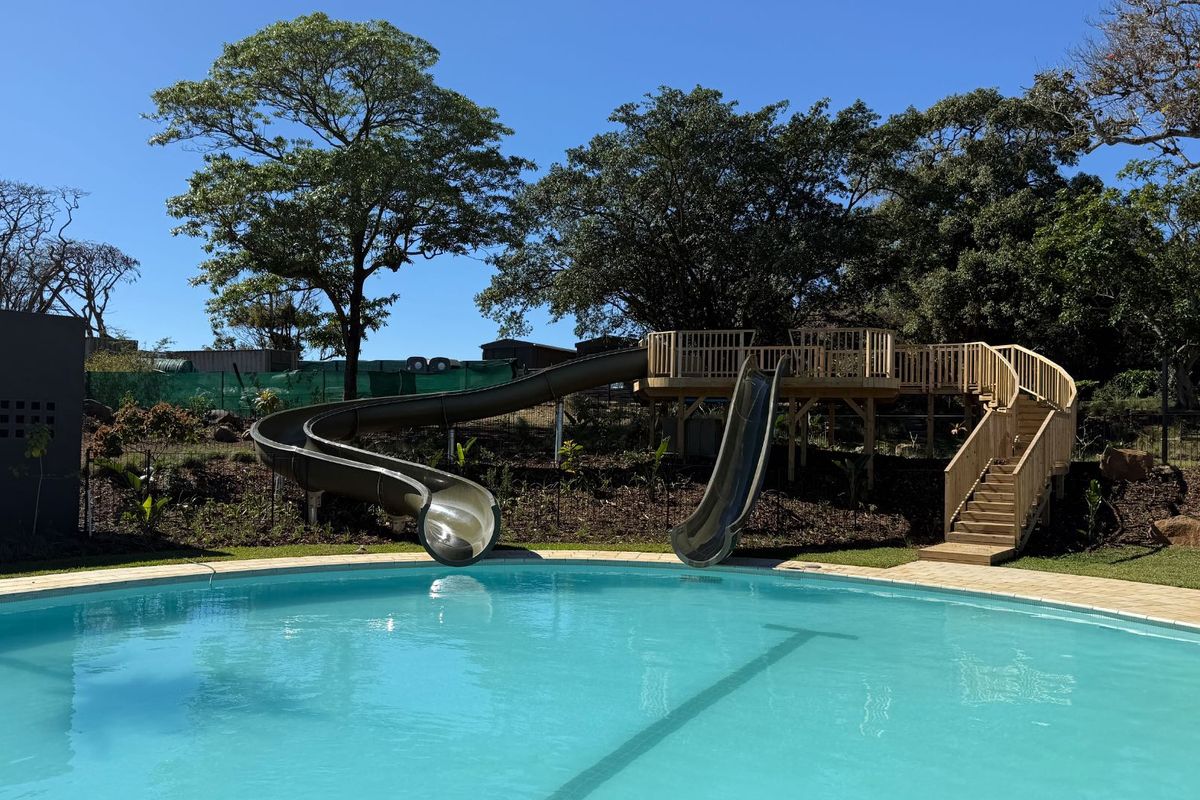 A well-maintained swimming pool area featuring a large pool, integrated water slides, and a wooden deck structure set within a landscaped garden.