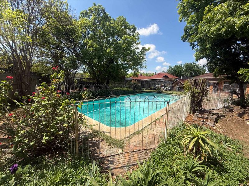 Outdoor swimming pool area featuring a paved surround and metal safety fencing, set within a mature garden environment.