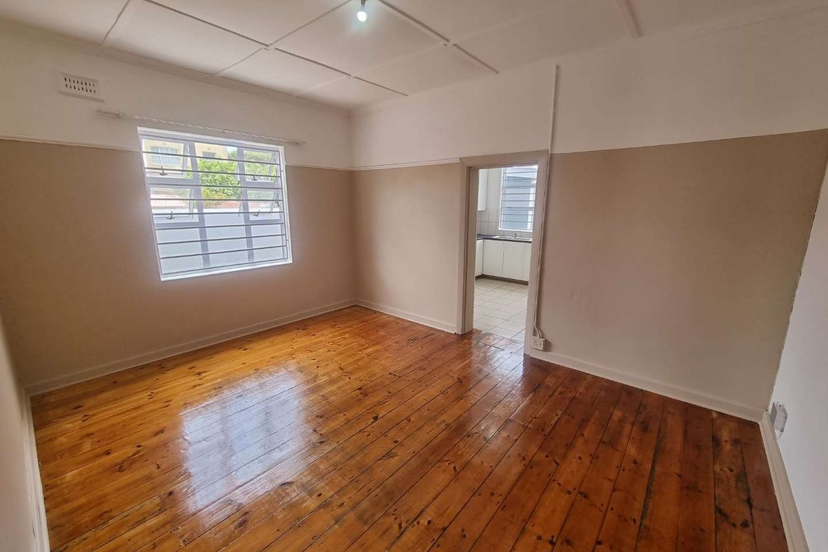 An empty living room featuring polished hardwood flooring, neutral two-tone walls, and a steel-framed window with security bars, leading into a kitchen area.