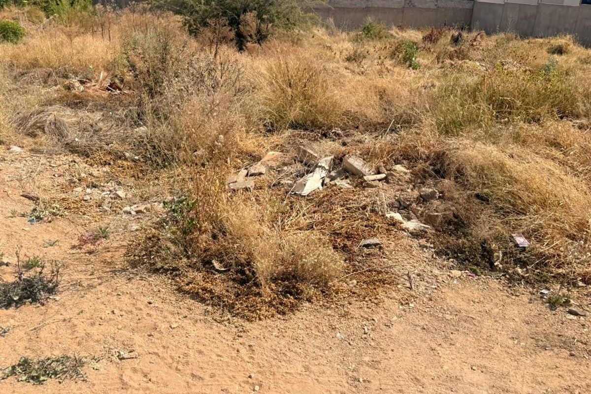 A view of the vacant residential land featuring dry, overgrown vegetation and scattered debris across the plot.
