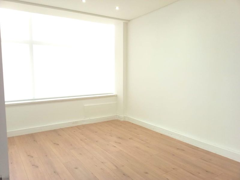 A bright, empty office space featuring modern laminate wood flooring, recessed ceiling lighting, and a large window providing ample natural light.