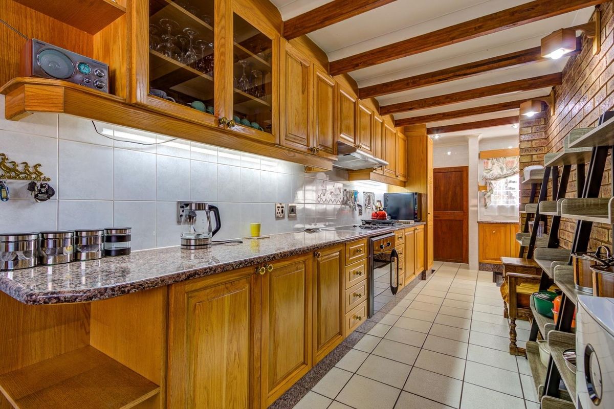 A functional kitchen featuring timber cabinetry, granite countertops, exposed timber ceiling beams, and a distinctive exposed brick wall with tiled flooring.