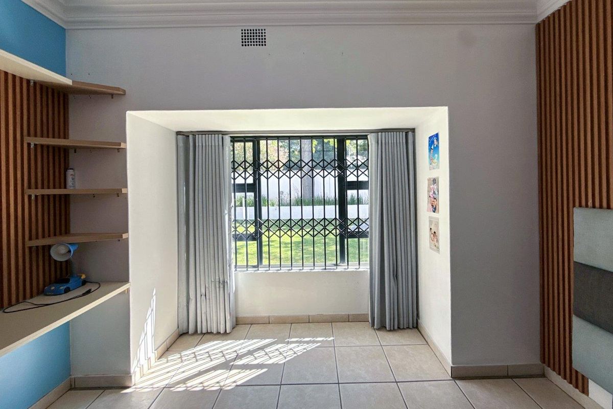 A bright bedroom featuring tiled flooring, built-in wooden shelving, decorative wall paneling, and a window secured with metal burglar bars.