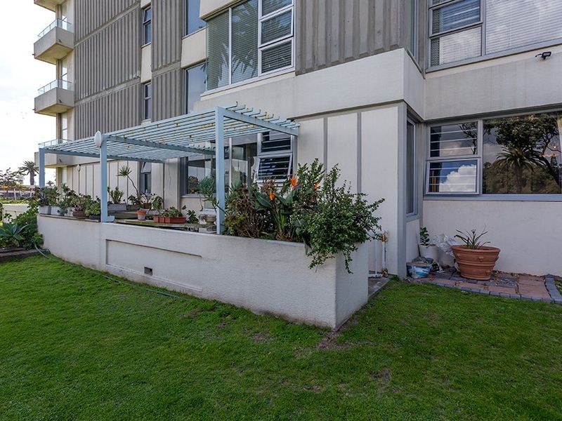 Rear exterior view of the apartment building featuring a concrete planter box, a metal pergola structure, and aluminium-framed windows overlooking a lawn area.
