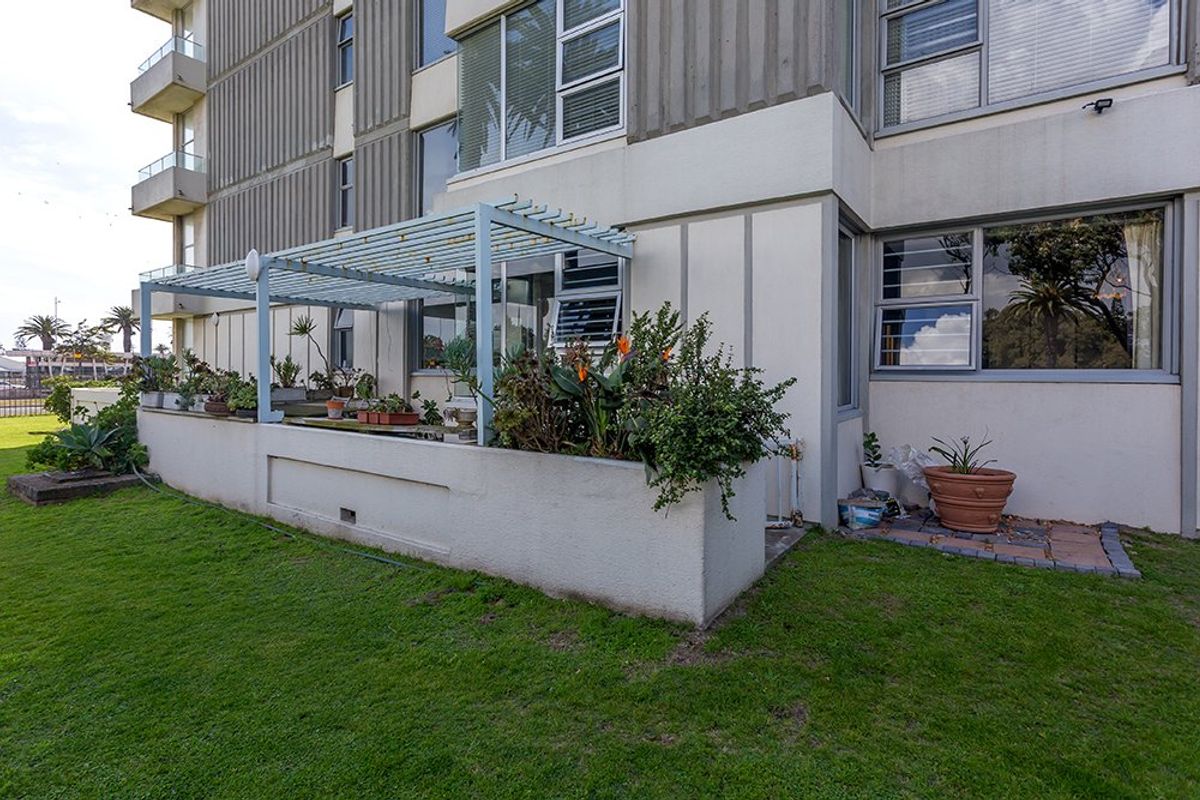 Rear exterior view of the apartment building featuring a concrete planter box, a metal pergola structure, and aluminium-framed windows overlooking a lawn area.