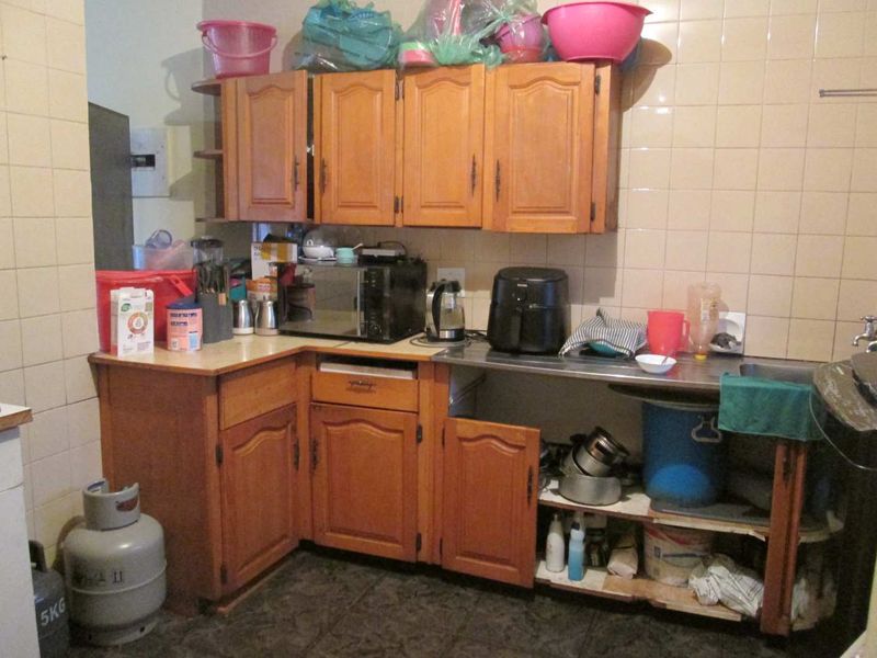 A dated kitchen featuring wooden cabinetry, tiled splashbacks, and a stainless steel sink, currently showing signs of wear and missing cabinet components.