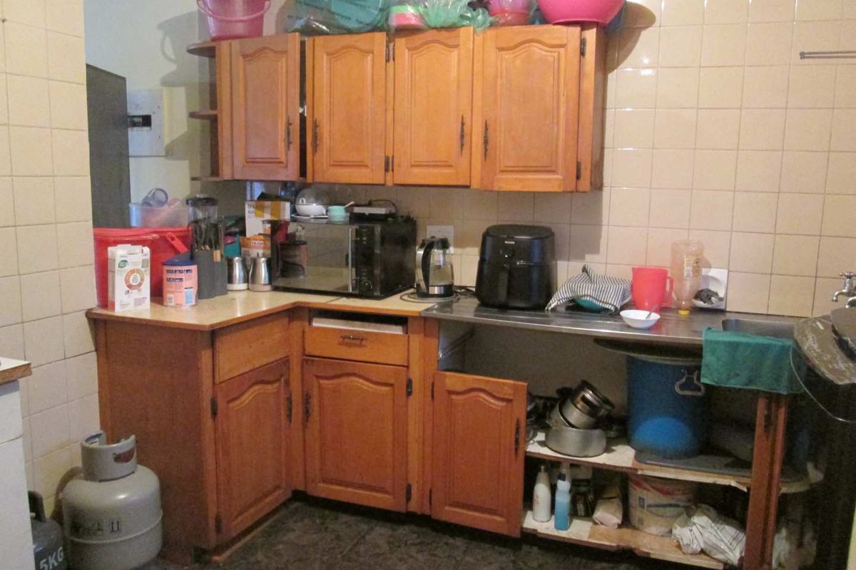 A dated kitchen featuring wooden cabinetry, tiled splashbacks, and a stainless steel sink, currently showing signs of wear and missing cabinet components.