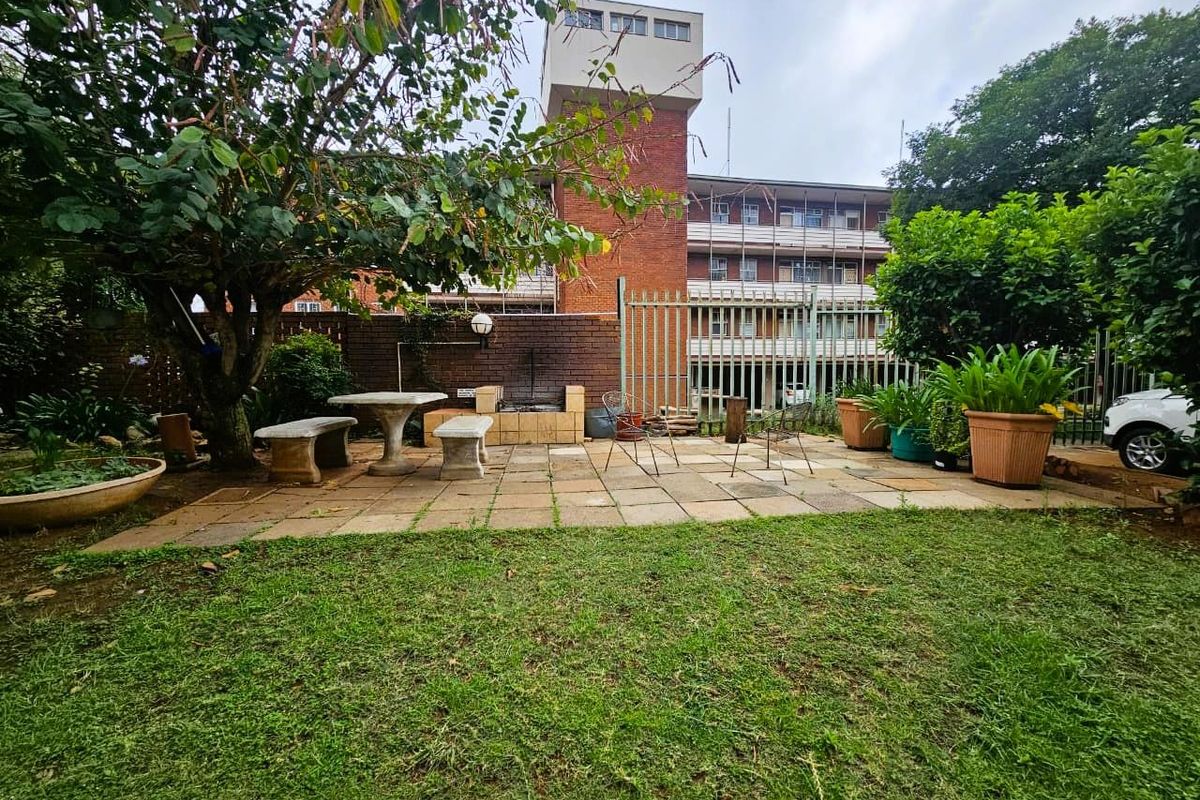A private rear garden area featuring a paved patio, built-in brick braai, and established trees, set against the backdrop of the apartment building.