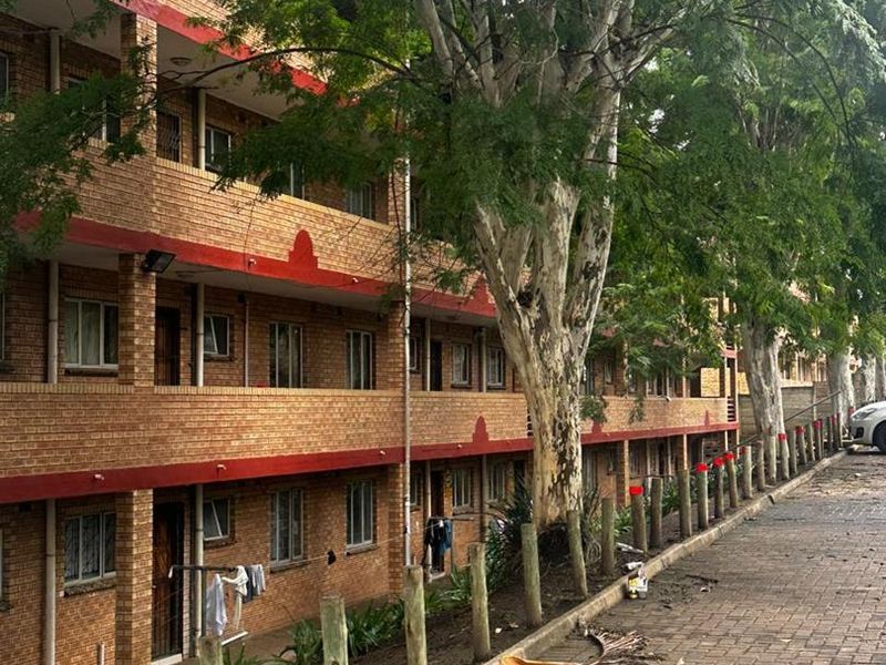 Exterior view of a multi-story brick apartment building featuring a paved driveway and mature trees along the front facade.